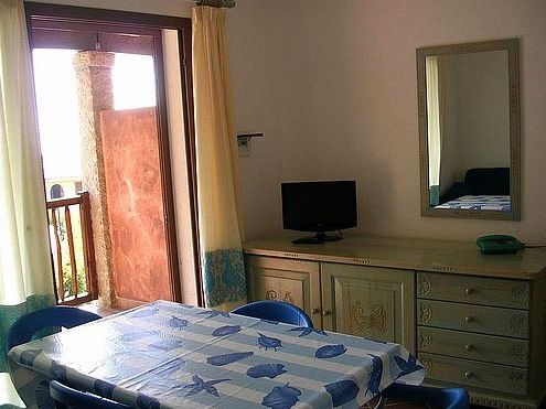 Sala con tavolo coperto, sedie blu, specchio e piccolo TV, porta aperta verso un balcone.