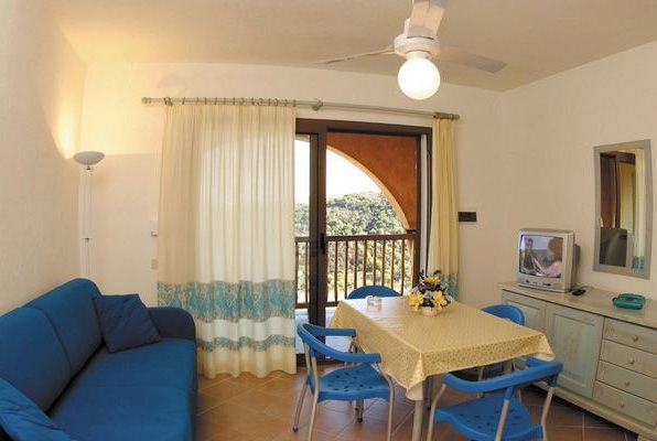Soggiorno con divano blu, tavolo con sedie, TV, ventilatore a soffitto, vista su un balcone.