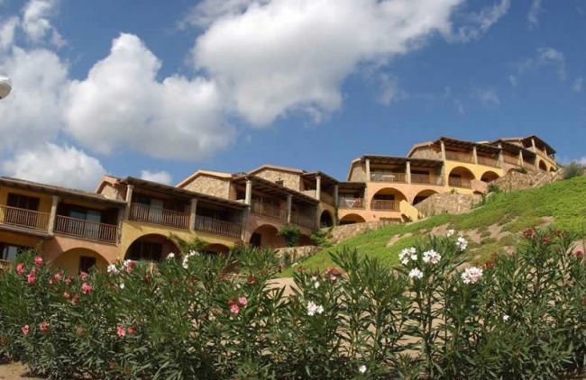 Edifici con archi e balconi su una collina con piante fiorite in primo piano.