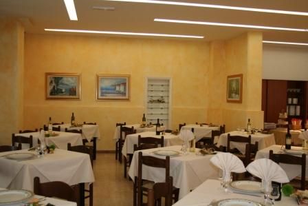 Ristorante con tavoli apparecchiati, tovaglie bianche, pareti giallo chiaro e quadri.