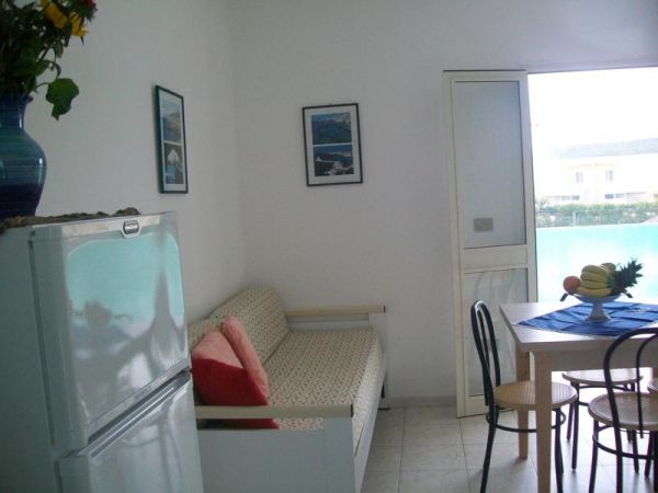 Soggiorno con divano, frigo e quadri, luce dal balcone.