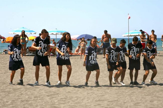 Linea di persone sulla spiaggia in divisa, sembrano eseguire una coreografia.