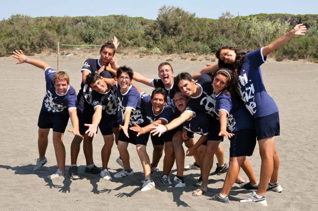 Un gruppo di persone in magliette blu posano sorridenti sulla spiaggia con le braccia aperte.