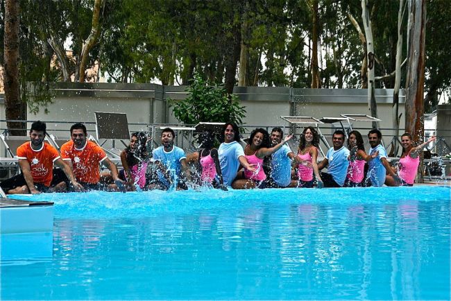 Persone in piscina che spruzzano acqua, sfondo con alberi.