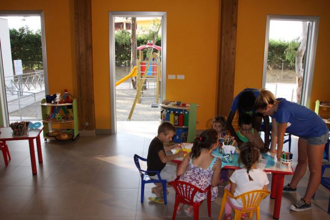 Bambini dipingono a un tavolo in un'aula colorata con finestre che mostrano un parco giochi.