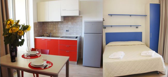 Interno di un appartamento con angolo cottura rosso e area notte con letto matrimoniale.