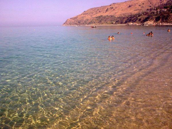Spiaggia con acqua cristallina e persone che nuotano, colline sullo sfondo.