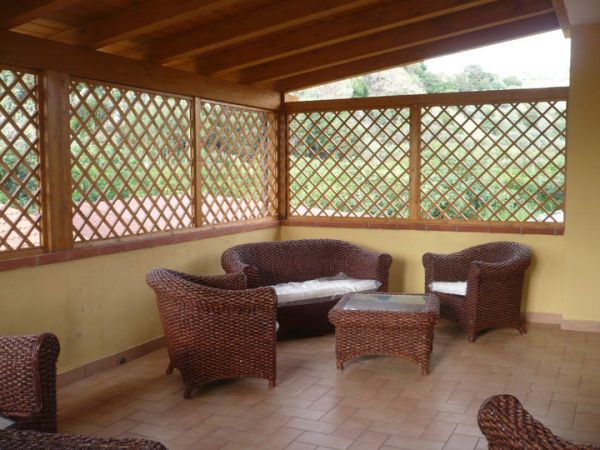 Veranda con mobili in vimini su pavimento in mattonelle, circondata da grigliati di legno.