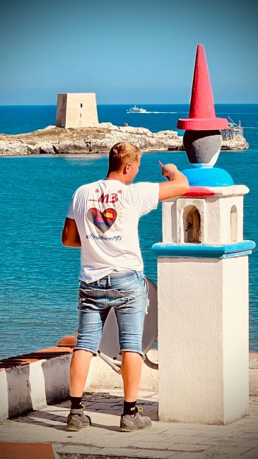 Un uomo dipinge una colonnina colorata vicino al mare, con una torre in lontananza.