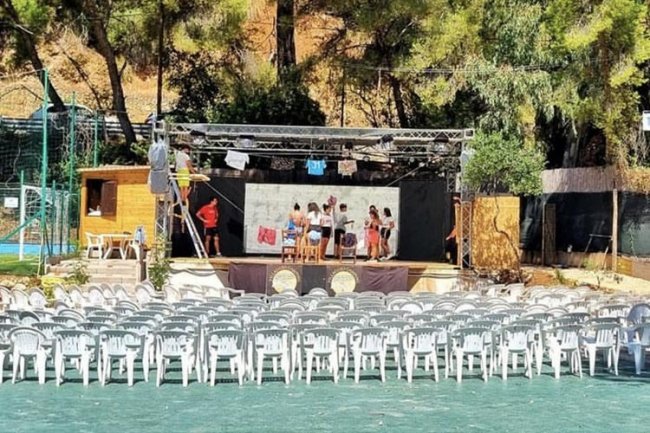 Teatro all’aperto con sedie bianche e palco.