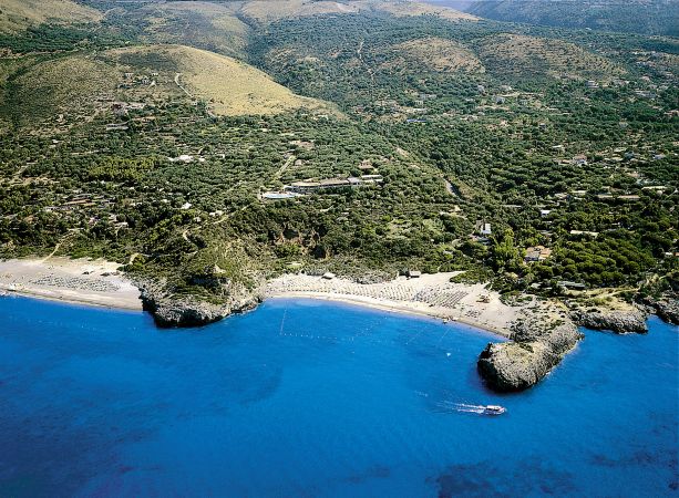 Una spiaggia curva con sabbia chiara, colline verdi e mare blu intenso.
