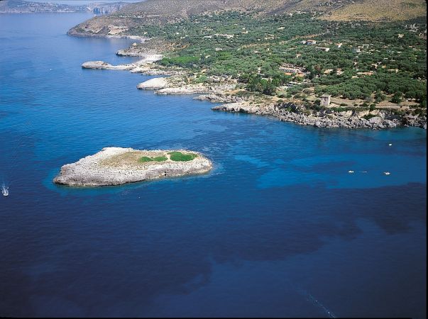 Costiera rocciosa con un'isoletta verde in mare, acqua trasparente e colline sullo sfondo.