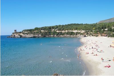 Spiaggia sabbiosa con bagnanti, mare calmo, una torre in lontananza e collina verde.