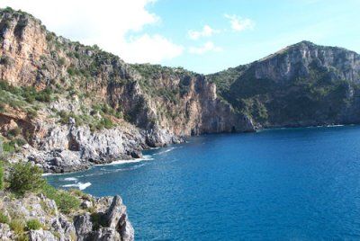 Scogliera rocciosa che si affaccia su un mare blu, con vegetazione sulla cima e cielo sereno.