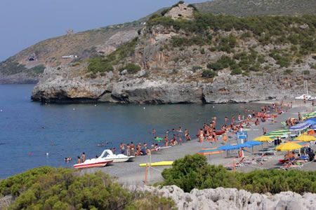 Spiaggia affollata con ombrelloni colorati, bagnanti in acqua e scogliere sullo sfondo.