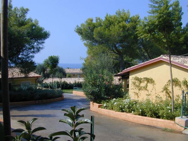 Un vialetto circondato da alberi e vegetazione, con edifici gialli e il mare visibile in lontananza.