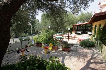 Spazio esterno di un ristorante con tavoli all'aperto, giostre per bambini e piante verdi.