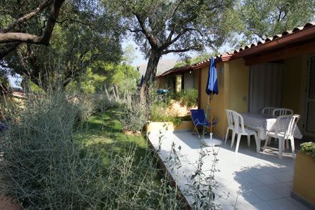 Terrazza di una casa con tavolo e sedie, circondata da cespugli e alberi.
