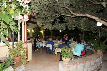 Persone sedute a tavoli all'aperto, circondate da piante, di sera.