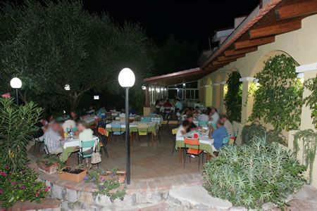 Cena all'aperto con persone sedute a tavoli sotto luci e accanto ad un edicola gialla.