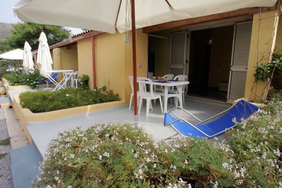 Piccolo patio con sedie bianche e una sdraio blu, protetto da grandi ombrelloni.