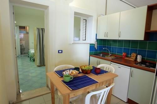 Angolo cucina con tavolo apparecchiato e porta verso zona notte con letto.