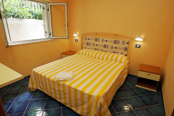 Camera con letto matrimoniale a righe gialle, pareti arancioni, pavimento blu e finestra aperta.