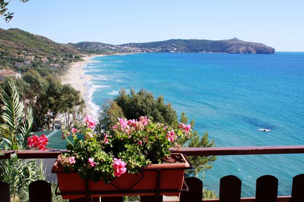 Un balcone con fiori rosa affacciato su una spiaggia con mare blu e colline in lontananza.