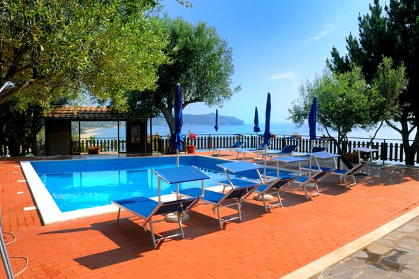 Piscina circondata da sedie a sdraio blu e ombrelloni, con vista mare e alberi.