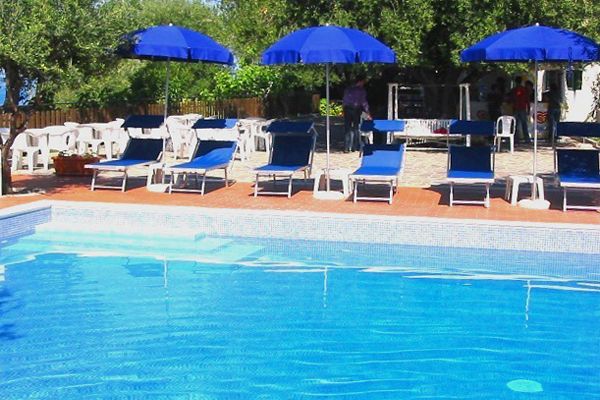Piscina con sedie a sdraio blu e ombrelloni, circondata da alberi verdi.