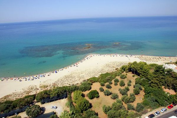 Una spiaggia di sabbia bianca con ombrelloni, mare turchese, alberi e un parcheggio vicino.