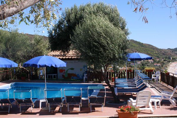 Una piscina circondata da lettini e ombrelloni blu, con alberi e paesaggio collinare sullo sfondo.