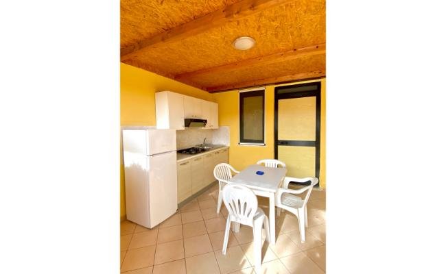 Cucina esterna con mobili bianchi, tavolo e sedie in plastica. Pareti gialle.