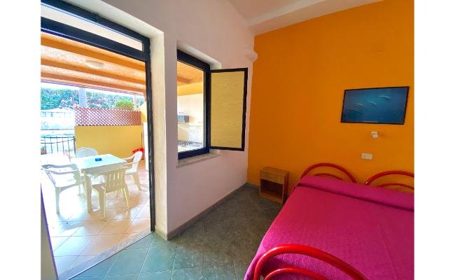 Stanza luminosa con pareti colorate, letto rosa e accesso ad un balcone con tavolo e sedie.