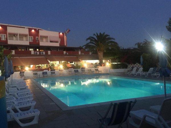 Piscina illuminata di notte, circondata da lettini, con palme e un edificio sullo sfondo.