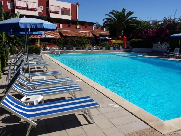 Piscina circondata da lettini blu a righe e ombrelloni, edificio sullo sfondo.