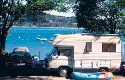 Un camper parcheggiato accanto a un lago con un gommone colorato e un'auto.