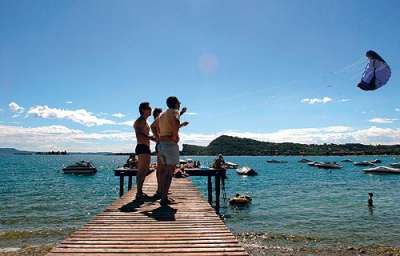 Persone su un molo di legno guardano un kite sul lago, con barche sullo sfondo.