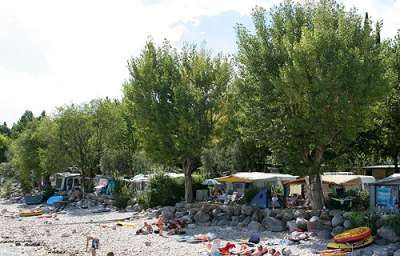 Una spiaggia di ciottoli con alberi, tende e gommoni colorati.