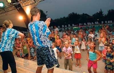 Due animatori in camicia hawaiana intrattengono un gruppo di bambini davanti a un palco.