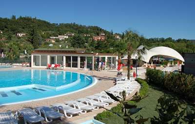 Una piscina contornata da lettini e ombrelloni, con un edificio basso e colline verdi sullo sfondo.
