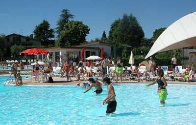Persone che giocano in una grande piscina all'aperto con sedie a sdraio e ombrelloni intorno.