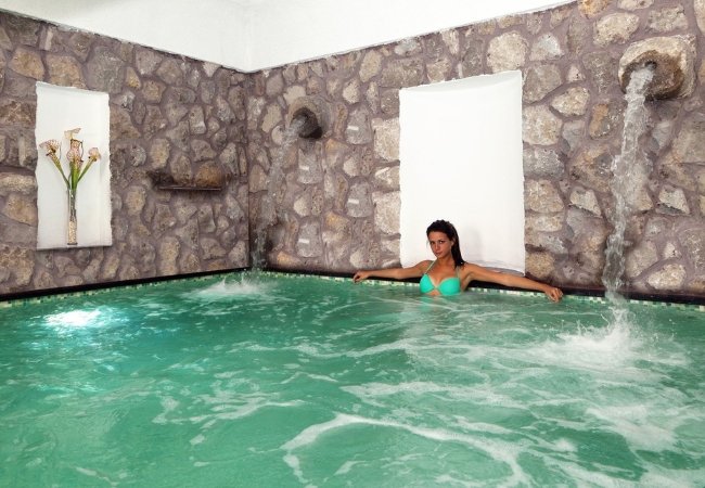 Una donna rilassata in piscina interna, circondata da pareti di pietra con cascate d'acqua.