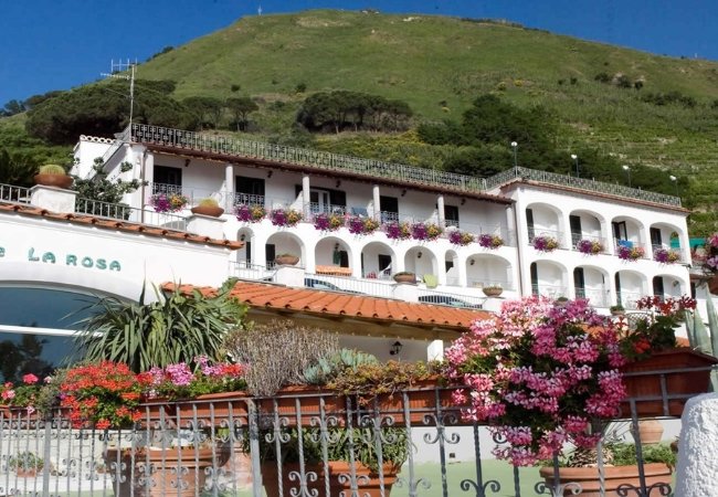 Edificio bianco con balconi e fiori, situato su una collina verdeggiante.