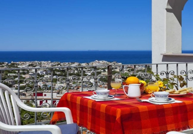 Tavolo all'aperto con tovaglia rossa, coppe e vista panoramica sul mare.