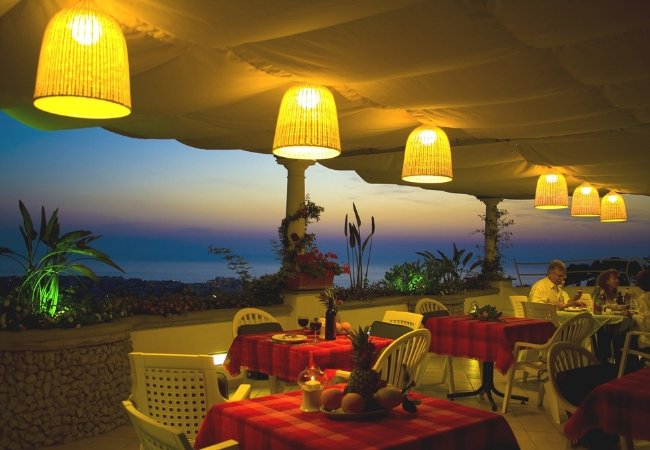 Ristorante all'aperto al tramonto con tavoli apparecchiati e lampade sospese.