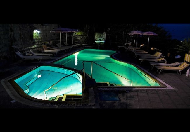 Piscina illuminata di notte con lettini e ombrelloni intorno.