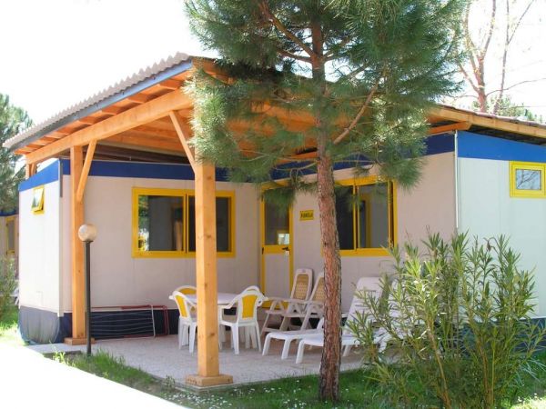 Bungalow con terrazza coperta, sedie bianche, albero e cespuglio in primo piano.