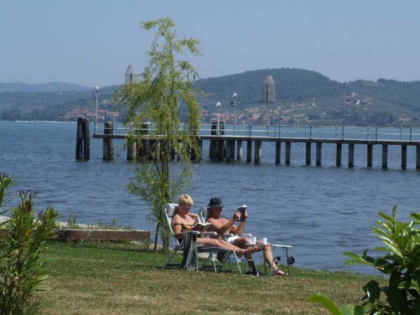 Due persone rilassate su sedie a sdraio vicino a un lago con un molo e colline sullo sfondo.