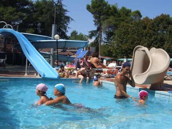 Una piscina con scivoli d'acqua, bambini che nuotano e persone che si rilassano.